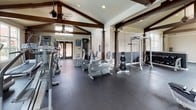 This fitness center is housed in a single-floor structure with striking architectural character. The vaulted ceiling, accented by bold exposed dark wood beam trusses, creates an open and airy atmosphere throughout the space. Recessed lighting and ceiling fans complement the abundant natural light that streams through multiple large windows, offering views of an outdoor pool area and surrounding greenery.

The facility is organized into distinct workout zones across a generously sized floor plan. The cardio area features a lineup of treadmills, elliptical trainers, and stationary bikes, all positioned to take advantage of the natural light and outdoor views. The strength training section is well-appointed with a cable machine, power rack, weight benches, and a full dumbbell rack with wall-mounted mirrors for form monitoring. Additional multi-use strength machines round out the equipment offerings.

Throughout the space, durable dark rubber flooring provides both safety and a clean, professional aesthetic. Decorative columns punctuate the interior, adding an architectural elegance that sets this facility apart from a standard gym. The overall ambiance is one of a well-maintained, thoughtfully designed fitness environment that balances functionality with refined style.