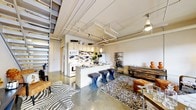 This three-story urban loft showcases an impressive industrial aesthetic defined by exposed ceiling pipes, concrete floors, and soaring ceiling heights that create a sense of openness and volume. The main living area is an open-concept space where the kitchen, dining, and lounge areas flow seamlessly together. The kitchen features light wood cabinetry, a subway tile backsplash, stainless steel appliances, and a central island with a granite countertop, complemented by stylish pendant lighting overhead. Bar-height stools line the island, making it an ideal casual dining or entertaining space. The living area is anchored by eclectic area rugs, including a bold zebra-print design, adding warmth to the cool concrete underfoot. A metal wine rack and reclaimed wood furniture lend a curated, industrial-chic character to the space. Industrial-style track and pendant lighting throughout enhances the loft's urban ambiance. A metal staircase connects the multiple floors, reinforcing the loft's raw architectural style. The space also includes a private balcony enclosed by a modern metal mesh railing, offering sweeping views of the surrounding urban skyline and cityscape below — a standout feature that brings the energy of the city directly into everyday living.