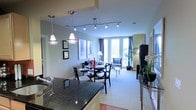 This well-appointed single-floor residence showcases an open-concept design that seamlessly connects the kitchen, dining, and living areas into one cohesive and functional space. The kitchen is anchored by a striking dark granite countertop that adds elegance and contrast, complemented by a stainless steel sink and modern pendant lighting that hangs gracefully overhead. Track lighting along the ceiling further enhances the kitchen and dining area, providing both style and practicality. The dining space comfortably accommodates a round glass-top table surrounded by contemporary black chairs, creating an intimate setting for meals. Beyond the dining area, the living room opens up generously, featuring neutral tones, plush carpeting, and tasteful furnishings. The space is flooded with natural light, courtesy of expansive floor-to-ceiling windows dressed with elegant drape curtains that frame views of the outdoors. Decorative framed artwork adorns the walls, and lush greenery adds a refreshing organic touch. The overall ambiance is modern yet inviting, with a sophisticated color palette of soft grays, greens, and warm wood tones that create a comfortable and refined living environment throughout.
