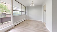 Located at 65 Park St E in Mississauga's Port Credit neighbourhood, this two-bedroom, one-bathroom unit offers a well-organized single-floor layout designed for comfortable everyday living. Light wood laminate flooring runs throughout the main living areas and bedrooms, creating a clean and cohesive aesthetic complemented by soft grey and white walls with crisp white baseboards and trim. The living room is generously proportioned, bathed in natural light from large floor-to-ceiling windows, and features a glass-panelled door that opens directly onto a private balcony with outdoor views. A chandelier-lit dining area adjacent to the kitchen further enhances the sense of openness. The kitchen is fitted with white cabinetry, a double sink, ample counter space, and light tile flooring, with a window bringing in additional natural light. The spacious primary bedroom features a dedicated closet and a window overlooking greenery, while the second bedroom is well-sized and equally bright with its own closet access. A hallway with an additional closet connects the rooms efficiently. The bathroom is finished with classic white subway tile surrounding a full soaker tub and shower combination. Overall, the unit presents a bright, clean, and functional living environment.