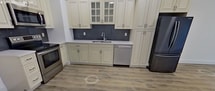 This 580-square-foot, one-bedroom, one-bathroom apartment on the second floor at 1070 River Rd in New Milford, NJ offers a clean, modern aesthetic throughout. The open-concept kitchen and living area is anchored by a well-appointed kitchen featuring cream-toned cabinetry with glass-front upper cabinets, a slate-blue tile backsplash, light quartz countertops, and a full suite of stainless steel appliances including a range, microwave, dishwasher, and French door refrigerator. Light wood-look luxury vinyl plank flooring flows consistently throughout all rooms, complementing the bright white walls and crisp white baseboards. The spacious 200-square-foot primary bedroom includes a wall-mounted PTAC climate control unit and windows that frame views of lush greenery. The bathroom features a full soaking tub with a shower, finished with classic white subway tile surround and chrome fixtures. A hallway connects the living spaces efficiently, contributing to the unit's practical and functional layout. Recessed lighting and natural light from multiple windows add to the apartment's clean, contemporary feel throughout.