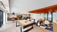 This contemporary amenity lounge is a thoughtfully designed social space that seamlessly blends recreation and relaxation. The single-floor layout is defined by an open floor plan with light hardwood flooring throughout, punctuated by bold design accents including a striking burnt-orange ceiling detail and coordinating accent walls that add warmth and visual interest to the space.

The recreation zone serves as a central focal point, featuring a full-size pool table and a foosball table positioned beneath sculptural black pendant lighting with gold interiors. A shuffleboard table extends the game options further. Floor-to-ceiling windows along multiple walls flood the space with natural light and offer expansive views of the surrounding cityscape and a courtyard pool area below.

Comfortable lounge seating is arranged throughout, including deep tufted sofas with accent pillows and an assortment of chairs set around a geometric marble-topped coffee table, all positioned atop bold patterned area rugs. A round dining-height table surrounded by curved black leather chairs provides a more structured gathering option.

A quieter lounge alcove at one end features patterned wallcovering, a sleek dark marble wet bar, and intimate seating arrangements separated by a floor-to-ceiling glass partition. Wall-mounted televisions and a built-in audio system complete the entertainment amenities, making this a fully equipped communal destination.