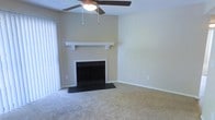 This single-story space centers around a comfortable living area designed with functional and classic elements. The room features a traditional fireplace framed by a dark surround and topped with a clean white mantel, serving as a natural focal point. Above, a ceiling fan with an integrated light fixture provides both air circulation and ambient lighting. Soft, wall-to-wall carpeting in a neutral tone covers the floor, contributing to a warm and understated atmosphere. The walls are finished in a light beige, creating a neutral backdrop that suits a variety of décor styles. Vertical blinds on one side of the room suggest access to natural light from an adjacent window or sliding door. The overall layout is open and straightforward, offering flexible furnishing options. Clean white baseboards trim the room, adding a crisp contrast to the neutral tones. The space presents a practical and inviting environment suitable for everyday living.