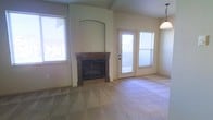 This single-story space centers around a comfortable living area defined by vaulted ceilings that lend a sense of openness and volume to the room. A standout feature is the built-in fireplace, framed by a warm wood mantel and set within a recessed arched niche, creating a natural focal point that adds character and architectural interest to the space. The fireplace surround extends to the ceiling, reinforcing the vertical emphasis of the room's design.

Soft, neutral-toned carpeting covers the floor throughout, contributing to a warm and inviting atmosphere. Multiple windows allow natural light to filter into the space, brightening the neutral wall tones and enhancing the overall sense of airiness. A glass-paneled door provides direct access to the outdoors, further connecting the interior to natural light and exterior views.

A pendant light fixture is partially visible, suggesting thoughtful overhead lighting to complement the natural illumination. The overall color palette — consisting of beige walls, warm wood tones, and light carpeting — creates a cohesive, understated aesthetic that offers a versatile backdrop for a variety of furnishing styles.