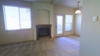 This single-story space centers around a comfortable living area defined by vaulted ceilings that lend a sense of openness and volume to the room. A standout feature is the built-in fireplace, framed by a warm wood mantel and set within a recessed arched niche, creating a natural focal point that adds character and architectural interest to the space. The fireplace surround extends to the ceiling, reinforcing the vertical emphasis of the room's design.

Soft, neutral-toned carpeting covers the floor throughout, contributing to a warm and inviting atmosphere. Multiple windows allow natural light to filter into the space, brightening the neutral wall tones and enhancing the overall sense of airiness. A glass-paneled door provides direct access to the outdoors, further connecting the interior to natural light and exterior views.

A pendant light fixture is partially visible, suggesting thoughtful overhead lighting to complement the natural illumination. The overall color palette — consisting of beige walls, warm wood tones, and light carpeting — creates a cohesive, understated aesthetic that offers a versatile backdrop for a variety of furnishing styles.