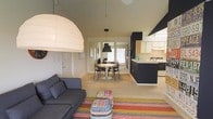 This single-floor space showcases an open-concept layout that integrates the living, dining, and kitchen areas into one cohesive environment. Vaulted ceilings amplify the sense of volume, while pendant lighting — including a large white dome fixture over the living area and a sleek black pendant above the dining space — adds purposeful illumination and visual interest.

The living area features a charcoal-toned sofa paired with a colorful striped area rug and a multicolored ottoman, lending warmth and personality to the neutral palette. Beige carpeting flows throughout the main areas, softening the overall aesthetic.

The dining zone centers around a round pedestal table surrounded by modern black wire-frame chairs, positioned near a window that introduces natural light. Adjacent to the dining area, the kitchen features dark navy cabinetry contrasted by light wood upper cabinets, a peninsula with counter seating, and integrated appliances. A bold dark hood anchors the kitchen visually.

The neutral wall tones, vaulted ceiling, and open sightlines throughout create a bright, airy ambiance that feels both functional and inviting.