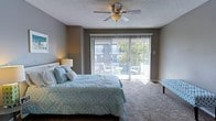 This two-floor space includes a well-appointed bedroom that serves as a restful retreat. The room features neutral grey walls and carpeted flooring that contribute to a calm, understated ambiance. A ceiling fan with an integrated light fixture is centrally mounted on the textured ceiling, providing both air circulation and ambient lighting. Natural light flows generously into the room through a large sliding glass door fitted with vertical blinds, which opens directly onto a private balcony. The balcony, visible through the glass, is furnished with seating and offers views of surrounding greenery, making it an inviting extension of the interior space. A bedside lamp adds warm, supplementary lighting to the room. The overall design is straightforward and functional, with a layout that maximizes usable floor space. The sliding door serves as a focal point, connecting the interior to the outdoors and enhancing the sense of openness. The space spans two floors, suggesting additional rooms and living areas beyond what is visible, offering a comfortable and practical multi-level living environment.