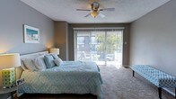 This two-floor space includes a well-appointed bedroom that serves as a restful retreat. The room features neutral grey walls and carpeted flooring that contribute to a calm, understated ambiance. A ceiling fan with an integrated light fixture is centrally mounted on the textured ceiling, providing both air circulation and ambient lighting. Natural light flows generously into the room through a large sliding glass door fitted with vertical blinds, which opens directly onto a private balcony. The balcony, visible through the glass, is furnished with seating and offers views of surrounding greenery, making it an inviting extension of the interior space. A bedside lamp adds warm, supplementary lighting to the room. The overall design is straightforward and functional, with a layout that maximizes usable floor space. The sliding door serves as a focal point, connecting the interior to the outdoors and enhancing the sense of openness. The space spans two floors, suggesting additional rooms and living areas beyond what is visible, offering a comfortable and practical multi-level living environment.