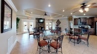 This single-story clubhouse offers a well-appointed communal gathering space designed with both comfort and functionality in mind. The main gathering room is a standout feature, showcasing a striking floor-to-ceiling stacked stone fireplace topped with a mounted flat-screen television, creating a warm and inviting focal point. Tray ceilings with crown molding add architectural elegance, while recessed lighting and a ceiling fan ensure year-round comfort. The space is furnished with ornate wrought iron bistro tables and chairs with cushioned seats, arranged throughout the room to accommodate multiple groups simultaneously. Large French doors line the perimeter, flooding the interior with natural light and providing direct access to an outdoor patio area. Light tile flooring extends throughout, contributing to the open, airy feel of the space. The kitchen area is fully equipped for event hosting, featuring rich dark wood cabinetry with raised panel detailing, granite countertops, stainless steel appliances including a range, refrigerator, dishwasher, and microwave, along with a coffee station. An arched window above the counter brings in additional natural light. The overall ambiance is warm, refined, and well-suited for community gatherings.