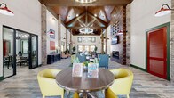 Located at 9500 Silver Collection Cir in Fredericksburg, VA, this single-story community clubhouse offers a generously sized, open-concept layout designed for both social gathering and private use. The centerpiece of the space is a dramatic vaulted ceiling clad in rich wood paneling, supported by exposed timber trusses and anchored by a statement chandelier that bathes the room in warm, inviting light. Towering natural stone columns frame the open floor plan, lending a rustic yet refined architectural character throughout.

The main lounge area is thoughtfully arranged with multiple seating vignettes, featuring a mix of vibrant yellow sofas, patterned armchairs, and casual round tables — creating flexible zones for relaxation and conversation. A prominent feature wall showcases a linear fireplace topped by a wall-mounted television, providing a cozy focal point. Red barn-style sconce lighting and green-trimmed windows and doorways add bold pops of color against the light wood-look tile flooring.

Adjacent seating nooks with striped and leather chairs offer quieter retreats, while a dedicated conference or leasing room provides privacy with large windows allowing abundant natural light. A kitchen or café area is also accessible from the main hall, rounding out this well-appointed community amenity space.
