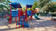 Located at 7761 Greenback Lane in Citrus Heights, California, this well-maintained outdoor community space offers a thoughtfully designed environment for recreation and relaxation. At its centerpiece is a colorful multi-level playground structure featuring bold red and blue tones, multiple climbing platforms, a spiral tube slide, interconnecting bridges, and educational panels — all set atop a generous wood chip safety surface that cushions the play area. The playground is fully enclosed by a sturdy wrought iron fence, providing a secure environment for children. Several green benches are positioned along the perimeter, offering comfortable seating for supervision and rest in the shade. Surrounding the playground, a spacious lawn of lush green turf provides additional open space for outdoor activities, bordered by elegant iron fencing and a paved walking path. Mature, large-canopy trees are dispersed throughout the grounds, casting natural shade across the space and contributing to a serene, park-like atmosphere. The residential buildings flanking the grounds feature a contemporary design with wood-tone siding, balconies, and well-tended landscaping with ornamental plantings. The overall environment is bright, open, and inviting, making it an ideal community gathering and play space.
