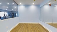 This single-floor studio space offers a clean, functional environment designed for movement and activity. The interior is defined by soft light blue walls that create a calm, open atmosphere, complemented by warm-toned wood-look laminate flooring that adds both durability and visual warmth underfoot. Recessed ceiling lighting provides even, well-distributed illumination throughout the space, ensuring a bright and welcoming setting at any time of day. Large wall-mounted mirrors are positioned strategically along the walls, enhancing the sense of spaciousness while serving a practical purpose for movement-based activities such as dance, yoga, fitness training, or similar pursuits. The white baseboard trim offers a crisp contrast against the blue walls, contributing to the overall polished and tidy aesthetic. The layout is open and unobstructed, maximizing usable floor space and allowing for flexible arrangement depending on the activity. A wall-mounted fixture, likely a speaker or sensor, adds a subtle functional element to the design. Overall, this studio presents a versatile, well-appointed space with a refreshing color palette and thoughtful design details suited to a wide range of instructional or recreational uses.