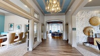 Located at 2951 Satellite Blvd in Duluth, Georgia, this single-floor clubhouse-style space offers a generously sized, thoughtfully designed interior that blends classic architectural details with contemporary décor. The hallway entry makes a striking first impression with light wood-look flooring, a geometric chandelier, and floor-to-ceiling framed mirror panels that create a sense of depth and openness. Arched doorways lead gracefully into adjoining areas, hinting at the sophisticated design throughout.

The expansive living room serves as the heart of the space, showcasing soaring ceilings adorned with rich, dark-stained exposed wood beams set against a vibrant teal ceiling — a bold design choice that adds warmth and personality. A carved stone fireplace with built-in cabinetry anchors one wall, while large windows flood the room with natural light and offer views of the surrounding greenery. Multiple seating arrangements — including plush sofas, accent chairs, and gold-finished side tables — create distinct yet cohesive conversation areas.

A cozy nook off the main area features a bright teal ceiling, white upholstered seating, blue patterned flooring, and honeycomb wall art, providing an intimate retreat within the larger space. Textured accent wallcovering, curated artwork, and layered area rugs complete the refined, welcoming ambiance throughout.