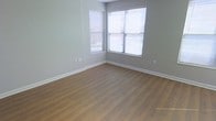 This single-floor space presents a clean, move-in-ready interior defined by a neutral and modern aesthetic. The room showcases warm-toned hardwood-style flooring that extends throughout, providing a cohesive and inviting foundation. The walls are finished in a soft gray tone, offering a versatile backdrop well-suited to a variety of furnishing styles.

A standout feature of the space is the corner window configuration, where two windows meet at an angle, allowing natural light to enter from multiple directions and brighten the room effectively. The windows are fitted with horizontal blinds, providing practical light control and privacy as needed. The surrounding views suggest an active neighborhood environment visible through the glass.

White baseboards cleanly frame the perimeter of the room, adding a crisp contrast against both the flooring and the gray walls. Electrical outlets are conveniently positioned along the walls. The overall condition of the space appears well-maintained, with no visible wear, making it an appealing and functional environment. The minimalist, unfurnished state of the room allows for a clear appreciation of its layout and natural light potential.