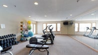 This single-floor amenity space is designed to support both active and leisure lifestyles, featuring two distinct areas: a fully equipped fitness center and a private movie screening room. The fitness center is bright and welcoming, with natural light streaming through multiple wood-trimmed windows set against neutral-toned walls. Recessed ceiling lighting supplements the daylight, illuminating a generous carpeted workout area. The room is outfitted with a variety of Precor cardio machines, including ellipticals and a treadmill, as well as a stationary bike and a weight bench. A multi-station cable machine and a rack of dumbbells support strength training, while a dedicated storage rack holds exercise balls, resistance bands, and foam rollers for functional fitness. A large floor-to-ceiling mirror along one wall enhances the sense of space, and wall-mounted televisions provide entertainment during workouts. The screening room offers a true cinema experience, featuring rows of plush, stadium-style seating in deep burgundy and black upholstery arranged on raised tiers for optimal viewing. A large flat-screen television mounted on a rich red accent wall serves as the focal point, while wood-trimmed windows and classic movie poster decor add character. Recessed lighting and neutral walls complete the inviting, theater-inspired atmosphere.