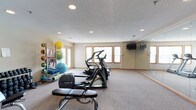 This single-floor amenity space is designed to support both active and leisure lifestyles, featuring two distinct areas: a fully equipped fitness center and a private movie screening room. The fitness center is bright and welcoming, with natural light streaming through multiple wood-trimmed windows set against neutral-toned walls. Recessed ceiling lighting supplements the daylight, illuminating a generous carpeted workout area. The room is outfitted with a variety of Precor cardio machines, including ellipticals and a treadmill, as well as a stationary bike and a weight bench. A multi-station cable machine and a rack of dumbbells support strength training, while a dedicated storage rack holds exercise balls, resistance bands, and foam rollers for functional fitness. A large floor-to-ceiling mirror along one wall enhances the sense of space, and wall-mounted televisions provide entertainment during workouts. The screening room offers a true cinema experience, featuring rows of plush, stadium-style seating in deep burgundy and black upholstery arranged on raised tiers for optimal viewing. A large flat-screen television mounted on a rich red accent wall serves as the focal point, while wood-trimmed windows and classic movie poster decor add character. Recessed lighting and neutral walls complete the inviting, theater-inspired atmosphere.