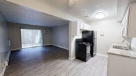 This well-appointed 1-bedroom, 1-bathroom unit offers a thoughtfully organized single-floor layout that maximizes every square foot. The open-concept design seamlessly connects the kitchen and living area, creating a cohesive and functional living space. The kitchen features a galley-style configuration with white cabinetry, light granite-style countertops, a stainless steel sink, and black appliances including a refrigerator, electric range, and dishwasher. Light wood-look flooring runs throughout the kitchen and living areas, while darker hardwood-style flooring adds contrast in the bedroom. The spacious living room is bathed in natural light through a sliding glass door that leads to a private enclosed outdoor patio area. A wall-mounted air conditioning unit and baseboard heating ensure year-round comfort. The bedroom is a well-sized, quiet retreat featuring a window with blinds offering outdoor views and continued baseboard heating. A hallway connects the main living spaces to the full bathroom, which includes a combined tub and shower, a single vanity, and matching light flooring. Textured ceilings and neutral gray-toned walls provide a clean, modern aesthetic throughout the unit.