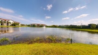 This single-story residence is set within a well-maintained community distinguished by its tranquil lakefront environment. The most prominent exterior feature is a expansive pond that stretches across the grounds, its calm surface animated by a central decorative fountain that adds a dynamic focal point to the landscape. Well-kept green lawns border the water's edge, creating a park-like atmosphere that enhances the overall sense of openness and natural beauty. Aquatic vegetation along the shoreline contributes to the setting's natural character. In the background, a Mediterranean-style multi-story residential building is visible, featuring a warm terracotta roofline and neutral facade framed by mature trees and dense tropical greenery — characteristic of Florida-style architecture. The surrounding landscape is bordered by tall trees that provide a natural screen, reinforcing a sense of privacy and seclusion. The open sky above allows abundant natural light to illuminate the grounds throughout the day. Interior images were not sufficiently lit to document specific room details; however, the exterior and community grounds present a peaceful, scenic living environment defined by its waterfront views and landscaped outdoor spaces.