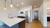 Located at 1633 N Arlington Pl in Milwaukee's vibrant 53202 zip code, this modern single-floor residence showcases contemporary design throughout. The open-concept layout seamlessly connects the kitchen and living area, creating a bright and functional space enhanced by light-toned luxury vinyl plank flooring. The kitchen is a standout feature, equipped with full stainless steel appliances including a French door refrigerator, electric range, and over-the-range microwave. Crisp white upper cabinetry contrasts elegantly with dark lower cabinets, while a white subway tile backsplash adds a classic touch. The large island with a quartz countertop and a distinctive blue base serves as both a functional workspace and a visual centerpiece, finished with a pendant light above. The spacious living area benefits from a large window that frames views of the surrounding Milwaukee neighborhood, flooding the room with natural light and hardwood-style flooring. The bedroom is comfortably carpeted and features a generously sized walk-in closet with built-in shelving, alongside a window overlooking mature trees. The bathroom is refined, with large-format tile flooring, a frameless glass walk-in shower with tile surround, a dark vanity with a white countertop, and a backlit mirror for an upscale touch.