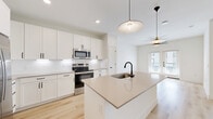 Located at 3200 Medical Center Drive in McKinney, TX, this thoughtfully designed one-bedroom, one-bathroom residence offers a clean, modern aesthetic with an efficient and functional layout. The open-concept living and kitchen area is anchored by a large center island with a quartz countertop, undermount sink, and matte black faucet, illuminated by stylish pendant lights. White shaker-style cabinetry reaches toward the ceiling, complemented by stainless steel appliances including a refrigerator with ice maker, electric range, and built-in microwave. Light-toned hardwood flooring flows throughout the main living spaces, enhancing the bright, airy atmosphere created by recessed lighting and natural light from glass panel doors. The bedroom is a comfortable retreat featuring plush carpet, a ceiling fan, and a window with outdoor views. It connects directly to a well-appointed bathroom with a quartz vanity, framed mirror, soaking tub, and tiled shower. A dedicated walk-in closet with built-in shelving and hanging rods provides generous storage. The overall design palette of crisp whites, warm wood tones, and black hardware accents lends the space a cohesive, contemporary feel.