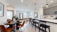 This single-floor residence showcases a thoughtfully designed open-concept layout that seamlessly connects the kitchen, dining, and living areas. Light wood-tone flooring flows throughout the main living spaces, complementing the neutral color palette and creating a bright, cohesive atmosphere.

The kitchen is a standout feature, equipped with stainless steel appliances, a combination of gray and light wood cabinetry, white quartz countertops, and a generously sized island with bar seating. Pendant lighting above the island adds a refined touch. The adjacent dining area accommodates a full table, while the living room benefits from natural light streaming through large windows and a glass-paneled door, with a ceiling fan overhead.

The two bedrooms are carpeted for comfort, each offering ample space and natural light. One bedroom connects directly to a well-designed en suite bathroom featuring a glass-enclosed walk-in shower, a double-sink vanity with light wood cabinetry, and white quartz countertops. A spacious walk-in closet with multi-level hanging rods and built-in shelving provides extensive storage.

Overall, the space delivers a clean, modern aesthetic with functional design elements throughout.