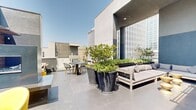 This two-floor space is defined by its impressive rooftop terraces, which together form a generous open-air environment with striking urban views of the surrounding cityscape. The outdoor areas are finished with large-format concrete pavers in a cool grey tone, lending a clean, contemporary aesthetic throughout.

Oversized dark planters housing mature trees and lush greenery serve as natural focal points, adding organic texture and privacy to the terraces. The upper level opens broadly to the sky, featuring a substantial seating arrangement with neutral-toned sectional sofas, low side tables with gold accents, and a dedicated dining area with a dark table and bistro-style chairs, all framed by an architectural concrete overhang that provides partial shade.

A lower terrace level offers a more intimate setting, incorporating warm horizontal wood-slatted screens that add texture against the dark blue-grey walls. A wall-mounted television is integrated into this feature wall, flanked by vibrant yellow accent chairs and additional lounge seating, making it suitable for both casual gatherings and outdoor entertainment. The interplay of natural light, curated greenery, varied seating zones, and city views creates a polished, versatile urban outdoor retreat.