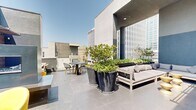 This two-floor space is defined by its impressive rooftop terraces, which together form a generous open-air environment with striking urban views of the surrounding cityscape. The outdoor areas are finished with large-format concrete pavers in a cool grey tone, lending a clean, contemporary aesthetic throughout.

Oversized dark planters housing mature trees and lush greenery serve as natural focal points, adding organic texture and privacy to the terraces. The upper level opens broadly to the sky, featuring a substantial seating arrangement with neutral-toned sectional sofas, low side tables with gold accents, and a dedicated dining area with a dark table and bistro-style chairs, all framed by an architectural concrete overhang that provides partial shade.

A lower terrace level offers a more intimate setting, incorporating warm horizontal wood-slatted screens that add texture against the dark blue-grey walls. A wall-mounted television is integrated into this feature wall, flanked by vibrant yellow accent chairs and additional lounge seating, making it suitable for both casual gatherings and outdoor entertainment. The interplay of natural light, curated greenery, varied seating zones, and city views creates a polished, versatile urban outdoor retreat.