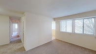 This single-floor residential unit offers a practical and straightforward layout across several well-defined rooms. The living area is generously proportioned, featuring neutral beige carpeting and clean white walls that create a bright, open atmosphere. A wide window fitted with horizontal blinds allows natural light to fill the space, lending it an airy quality. A partial wall divides the living area, adding subtle definition without compromising the sense of openness.

The bedrooms follow a similar design, each finished with wall-to-wall carpet and windows that provide outdoor views and natural light. The rooms are simply appointed, offering a blank canvas adaptable to various arrangements.

The galley-style kitchen is efficiently organized, featuring warm-toned wooden cabinetry both above and below the countertops, laminate counters with an integrated sink, and tile flooring. White appliances, including a refrigerator and range, are included. A flush ceiling light provides functional illumination throughout the kitchen corridor.

Two bathrooms serve the unit, each equipped with a vanity, mirror, and toilet. One bathroom features a bright vanity light strip above a medicine cabinet mirror, while the other includes a towel bar and tiled shower surround. Both are finished in neutral tones, maintaining the consistent, understated aesthetic found throughout the unit.