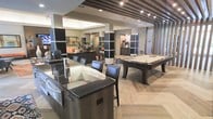 This single-floor amenity space is designed with a contemporary aesthetic, offering a seamlessly connected layout that blends entertainment and relaxation. The space features a striking wet bar or game table island with a dark granite countertop and built-in lighting, accompanied by sleek leather bar-height chairs — ideal for social gatherings. Adjacent to this central hub sits a full-sized billiards table set atop elegant herringbone-patterned hardwood-style flooring, providing a sophisticated focal point for recreation. Structural columns with dark wood paneling add architectural interest while defining separate activity zones without fully enclosing them. The ceiling draws the eye upward with a distinctive slatted wood detail, adding warmth and visual depth to the space. Beyond the game area, a well-appointed lounge zone offers plush seating, accent pillows, and a mounted flat-screen television, creating a cozy retreat for relaxing. Recessed lighting throughout ensures the entire space is well-lit, while decorative accents and stone-clad column details contribute to the upscale, modern ambiance. The open, flowing floor plan makes this space equally suited for casual everyday use and larger social events.