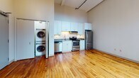 Located at 95 Chestnut Street in the heart of Providence, Rhode Island, this single-floor loft unit showcases a compelling blend of historic industrial architecture and contemporary updates. Original hardwood floors with rich, warm tones run throughout the open living space, reflecting the building's storied past. Exposed brick walls — some left in their natural red state, others painted white — add textural depth and character, while arched windows with black frames allow generous natural light to pour into the space and offer views of the surrounding urban streetscape.

The soaring ceilings create an airy, expansive atmosphere, accentuated by visible ductwork and industrial-style pendant lighting that nod to the building's heritage. The kitchen is thoughtfully appointed with white shaker cabinetry, dark stone countertops, stainless steel appliances including a refrigerator, dishwasher, and range, and an under-cabinet range hood. An in-unit stacked washer and dryer is conveniently tucked behind a glass-panel sliding door adjacent to the kitchen.

The bathroom features clean, modern finishes including white subway tile surrounding the tub, light tile flooring, a white vanity with a stone-topped sink, and a mirrored medicine cabinet. The overall ambiance balances raw, historic charm with functional, updated living amenities.