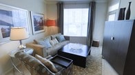 This single-floor space centers around a thoughtfully designed living area that balances comfort and style. The sitting area is anchored by a plush, neutral-toned sofa adorned with decorative teal and patterned throw pillows, complemented by a matching upholstered chair. A dark espresso square coffee table with a built-in lower shelf serves as the room's focal point, topped with a decorative crystal bowl that adds an elegant touch. A textured area rug in warm tones grounds the seating arrangement and adds visual warmth to the space. Natural light filters generously through a large central window dressed with classic horizontal blinds and floor-length charcoal drape panels, creating a bright yet intimate atmosphere. A striking orange tripod floor lamp provides warm ambient lighting, while a framed cityscape artwork on the wall adds a metropolitan character to the room. The neutral wall tones throughout enhance the sense of openness. Overall, the space presents a cohesive, contemporary aesthetic that is both functional and inviting.