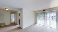 This single-floor residence offers a clean, functional layout designed for comfortable everyday living. The main living area is finished with wall-to-wall carpeting in a neutral tone, creating a warm and inviting atmosphere throughout the space. A large sliding glass door with vertical blinds fills the room with natural light while providing direct access to an outdoor area, where views of surrounding greenery can be enjoyed.

The living space flows openly toward a partially partitioned area, where a structural dividing wall creates a subtle separation between the main room and the kitchen beyond. The kitchen is visible through the opening and features dark wood cabinetry, offering a practical contrast to the light-toned walls and flooring found throughout the rest of the space. A hallway extends further into the residence, hinting at additional rooms and providing a sense of depth to the overall floor plan.

Crisp white walls and a smooth ceiling with integrated HVAC venting contribute to the space's clean, understated aesthetic. The neutral color palette throughout creates a versatile backdrop that adapts easily to a variety of interior styles. Overall, the space presents a straightforward and practical single-level layout with a comfortable connection between interior living areas and the outdoors.