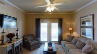 This single-floor living space features a warm and inviting design highlighted by golden-tan walls that create a cozy, intimate atmosphere. Crown molding runs along the ceiling perimeter, adding a classic architectural detail that enhances the room's refined character. A white ceiling fan with an integrated light fixture serves as a central focal point overhead, providing both air circulation and ambient lighting.

Natural light enters through a pair of windows dressed with dark curtain panels, which offer contrast against the warm wall tones while allowing adjustable light control via fitted blinds. A floor lamp positioned in the corner contributes additional warm, accent lighting to the space.

The living area is thoughtfully arranged with a striped upholstered armchair and a cushioned sofa, both offering comfortable seating. A rectangular wooden coffee table anchors the seating arrangement atop a traditional area rug with floral detailing, grounding the layout and adding texture to the light-toned carpeted floor. The overall ambiance is relaxed and traditional, with a balanced mix of functional furnishings and decorative elements that make the space feel well-appointed and livable.