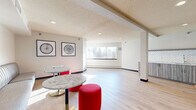This single-floor space is thoughtfully designed as a communal lounge or common area, offering an open, flexible layout across a compact yet functional footprint. The living area is the heart of the space, featuring light-toned hardwood flooring that extends throughout, creating a clean and cohesive aesthetic. Recessed lighting and a textured ceiling complement the bright, neutral white walls, while large windows along one side of the room allow natural light to fill the interior and offer views of surrounding greenery.

The seating arrangement is varied and inviting, incorporating a tufted gray banquette paired with marble-topped bistro tables, bold red upholstered ottomans, mid-century modern leather armchairs, and additional accent seating. Framed black-and-white artwork decorates the walls, adding visual interest throughout. A kitchenette area along one wall features gray cabinetry with a herringbone tile backsplash and a sink, providing practical utility for the space.

The bathroom is accessible and well-appointed, equipped with grab bars, a toilet, and a vanity, finished with the same light hardwood flooring. Dark-toned doors throughout provide a modern contrast to the bright interior. Overall, the space presents a welcoming, contemporary atmosphere well-suited for social gathering or collaborative use.