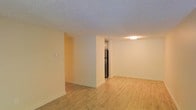 This single-floor space offers a clean, functional layout designed to maximize openness and usability. The main living area is finished with light-toned hardwood flooring that runs throughout, lending warmth and a sense of continuity to the space. Walls are painted in a neutral cream tone, creating a bright and welcoming atmosphere that pairs well with the natural light filtering through the room.

A partial dividing wall separates the living area from the kitchen zone, providing a degree of definition between spaces while maintaining an open feel. The kitchen is positioned just beyond this partition, where a stainless steel refrigerator is visible, hinting at modern appliances within. Overhead lighting in the kitchen area ensures the space remains well-lit regardless of natural light conditions.

The textured popcorn ceiling adds a classic character to the interior, complementing the simple, understated design of the space. White baseboards frame the flooring throughout, adding a clean, finished look to each room. The layout is straightforward and efficient, making it well-suited for a variety of uses. Overall, the space presents a practical and inviting environment with a neutral palette that allows for flexible interior styling.