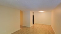This single-floor space offers a clean, functional layout designed to maximize openness and usability. The main living area is finished with light-toned hardwood flooring that runs throughout, lending warmth and a sense of continuity to the space. Walls are painted in a neutral cream tone, creating a bright and welcoming atmosphere that pairs well with the natural light filtering through the room.

A partial dividing wall separates the living area from the kitchen zone, providing a degree of definition between spaces while maintaining an open feel. The kitchen is positioned just beyond this partition, where a stainless steel refrigerator is visible, hinting at modern appliances within. Overhead lighting in the kitchen area ensures the space remains well-lit regardless of natural light conditions.

The textured popcorn ceiling adds a classic character to the interior, complementing the simple, understated design of the space. White baseboards frame the flooring throughout, adding a clean, finished look to each room. The layout is straightforward and efficient, making it well-suited for a variety of uses. Overall, the space presents a practical and inviting environment with a neutral palette that allows for flexible interior styling.