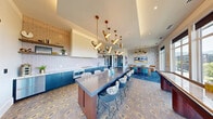 Located at 357 E 500 S in Salt Lake City, Utah, this thoughtfully designed amenity lounge offers a compelling blend of modern aesthetics and functional versatility. The space unfolds across a single floor, encompassing a fully equipped kitchen, an open social area, and a private workstation nook.

The kitchen is a standout feature, showcasing bold teal cabinetry paired with marble-look countertops, a patterned tile backsplash, open wood shelving, and stainless steel appliances. A large central island with a dark concrete-style surface provides generous bar seating, anchored by a dramatic sculptural pendant light installation in brushed gold that draws the eye upward.

The adjoining lounge and dining area continues the vibrant design language, with deep blue patterned flooring, an intricate floral tile floor, bold wallcoverings, and additional statement pendant lighting. A shuffleboard table adds a recreational element, while floor-to-ceiling windows flood the space with natural light and frame expansive views of the Salt Lake City skyline, surrounding greenery, and the distant Wasatch Mountains.

A private workstation alcove offers a quiet retreat, outfitted with a built-in floating desk, a plush velvet chair, acoustic wall panels, and elegant wall-mounted lighting — ideal for focused work or private meetings. Together, these areas create a dynamic, design-forward environment suited for both social gatherings and productive use.