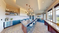 Located at 357 E 500 S in Salt Lake City, Utah, this thoughtfully designed amenity lounge offers a compelling blend of modern aesthetics and functional versatility. The space unfolds across a single floor, encompassing a fully equipped kitchen, an open social area, and a private workstation nook.

The kitchen is a standout feature, showcasing bold teal cabinetry paired with marble-look countertops, a patterned tile backsplash, open wood shelving, and stainless steel appliances. A large central island with a dark concrete-style surface provides generous bar seating, anchored by a dramatic sculptural pendant light installation in brushed gold that draws the eye upward.

The adjoining lounge and dining area continues the vibrant design language, with deep blue patterned flooring, an intricate floral tile floor, bold wallcoverings, and additional statement pendant lighting. A shuffleboard table adds a recreational element, while floor-to-ceiling windows flood the space with natural light and frame expansive views of the Salt Lake City skyline, surrounding greenery, and the distant Wasatch Mountains.

A private workstation alcove offers a quiet retreat, outfitted with a built-in floating desk, a plush velvet chair, acoustic wall panels, and elegant wall-mounted lighting — ideal for focused work or private meetings. Together, these areas create a dynamic, design-forward environment suited for both social gatherings and productive use.