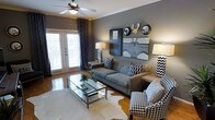 This living space showcases a thoughtfully curated interior with a sophisticated gray and black-and-white color scheme throughout. The room centers around a gray tufted sofa adorned with geometric and diamond-patterned throw pillows, complemented by a bold houndstooth accent chair that adds visual interest. A glass-and-metal coffee table anchors the seating arrangement atop a cowhide rug, which adds a subtle organic contrast to the otherwise modern aesthetic. Crown molding frames the ceiling, where a classic ceiling fan with a light fixture provides both comfort and illumination. The focal wall features a large Brooklyn Bridge canvas print flanked by decorative circular mirrors and a sculptural moose head accent, creating a striking gallery-style display. Warm table lamps with geometric black-and-white bases flank the seating area, providing layered ambient lighting. French doors draped with dark patterned curtains flood the space with natural light while offering direct access to an outdoor area. Hardwood flooring runs throughout, adding warmth to the cool-toned palette and tying the overall design together cohesively.