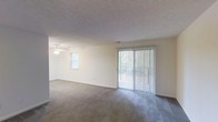 This single-floor space offers a clean, open layout designed for comfortable everyday living. The main living area is generously proportioned, featuring soft wall-to-wall carpeting that adds warmth underfoot. A textured ceiling runs throughout, lending a classic residential feel to the interior. Natural light plays a prominent role in the ambiance of this space — a side window and a full-width sliding glass door fitted with vertical blinds work together to brighten the room throughout the day while offering flexible light control and outdoor access. The sliding glass door serves as a focal point along the main wall, providing a seamless visual connection to the exterior. A ceiling fan with a light fixture is positioned overhead, ensuring year-round comfort and additional illumination. The neutral color palette of soft white walls and light-toned carpet creates a versatile backdrop, making the space easy to personalize and furnish. Overall, the layout is practical and inviting, with an airy atmosphere well-suited for a variety of living arrangements.