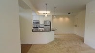 This single-floor residence offers a thoughtfully designed open-concept layout that seamlessly connects the kitchen and living areas. The kitchen is a standout feature, equipped with stainless steel appliances including a refrigerator, electric range, and microwave. Rich dark granite countertops curve elegantly into a breakfast bar, providing a natural division between the kitchen and the carpeted living space. White cabinetry lends a clean, classic look, while two pendant lights hang above the bar, adding a warm, welcoming ambiance. Recessed track lighting along the ceiling of the living area further enhances the bright, open feel of the space. The living area is finished with plush neutral carpeting, creating a comfortable and cohesive atmosphere. The transition between the kitchen's hardwood-style flooring and the carpeted living room adds a subtle distinction between the functional and relaxing zones of the home. Light, neutral wall tones throughout contribute to an airy and inviting environment. Ceiling vents are thoughtfully integrated to maintain a clean architectural finish. Overall, the space strikes a balance between functionality and comfort, making it ideal for both everyday living and casual entertaining.