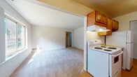 This single-floor residence offers a practical and comfortable layout across two bedrooms and one full bathroom. The open-concept living area is carpeted and connects seamlessly to the kitchen, creating a cohesive and functional common space. A large picture window in the living area allows natural light to filter in, providing views of the surrounding greenery outdoors. A ceiling fan overhead adds to the room's functionality.

The galley-style kitchen features warm-toned wood cabinetry lining both walls, offering generous upper and lower storage. White electric range, refrigerator, and a sink with laminate countertops complete the kitchen's practical setup. Hardwood-style flooring runs throughout the kitchen, contrasting with the carpeted living spaces.

Both bedrooms are carpeted and generously proportioned, each featuring windows that bring in natural light. One bedroom includes a walk-in closet with built-in shelving, while the other provides closet access and multiple doorways for flexible use.

The full bathroom is clean and simply appointed, featuring a tiled tub-shower combination and tile flooring. The overall ambiance throughout is neutral and move-in ready, with a straightforward layout well-suited for everyday living.