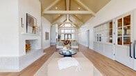 Located at 23615 Kingsland Blvd in Houston, TX, this single-floor space offers a thoughtfully designed interior that seamlessly blends warmth, elegance, and functionality across a generously sized layout.

The living room is the architectural centerpiece of the space, showcasing a soaring vaulted ceiling with exposed wood beams and tongue-and-groove detailing that draws the eye upward. Wide-plank oak hardwood floors run throughout, while a tiled fireplace serves as a striking focal point. Floor-to-ceiling windows and glass doors flood the room with abundant natural light and provide open views of the surrounding outdoors. Graceful arched passageways connect the living area to adjacent spaces, reinforcing a cohesive and airy design language.

The dining room exudes refined elegance, featuring stone tile flooring, dramatic floor-to-ceiling black-framed windows dressed with linen drapery, and an intricate botanical wallcovering that adds texture and depth. A substantial wood dining table surrounded by woven rattan chairs anchors the room, while a classic wood credenza completes the sophisticated setting.

The kitchen area features a large waterfall-edge marble island with seating, warm wood cabinetry, marble tile backsplash, and pendant lighting beneath an exposed wood ceiling beam. Throughout the space, a neutral palette of whites, natural woods, and earthy tones creates a serene, cohesive ambiance.