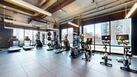 Located at 180 Morgan St in Jersey City, NJ, this modern fitness center offers a comprehensive exercise environment designed to accommodate a wide range of training styles. The space features an industrial-chic aesthetic, with exposed ductwork and piping running along the ceiling, complemented by sleek linear lighting and a dark, sophisticated color palette accented by warm wood-paneled walls.

The cardio zone is well-appointed with multiple treadmills, elliptical trainers, and stationary bikes, all positioned along floor-to-ceiling windows that flood the space with natural light and frame dynamic views of the surrounding Jersey City skyline. A dedicated strength training area houses an extensive free weight selection, weight benches, and cable machines, with large mirrors lining the walls to maximize the sense of openness.

A functional training zone features TRX suspension equipment and a multi-use rig, ideal for bodyweight and resistance training. Stability balls and kettlebells are neatly organized throughout. An additional open studio area with hardwood flooring and mirrored walls provides a versatile space for stretching, yoga, or group fitness. Large windows throughout offer views of an adjacent outdoor green space, enhancing the overall bright and energizing atmosphere of this well-rounded facility.