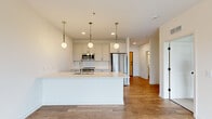 This well-appointed single-floor unit offers a thoughtfully designed layout encompassing one bedroom, one bathroom, a combined kitchen and dining area, a dedicated laundry room, and a closet. The open-concept kitchen is a standout feature, centered around a large white quartz island with an integrated sink and pull-down faucet, providing ample prep space and casual seating. White shaker-style cabinetry extends to the ceiling, complemented by a geometric tile backsplash and a full suite of stainless steel appliances, including a refrigerator, microwave, and range. Three globe pendant lights with brushed metal fixtures hang above the island, adding a contemporary touch. Warm hardwood-style flooring flows throughout the main living areas, while the bedroom features plush carpeting and a large picture window that frames tree-lined neighborhood views, filling the space with natural light. A private balcony is accessible from the living area. The bathroom includes a full soaking tub with clean white tile surround. The laundry room houses a stacked washer and dryer unit alongside a water heater. Clean white walls and recessed lighting throughout contribute to a bright, modern ambiance.
