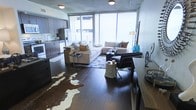 This single-floor space showcases a thoughtfully designed open-concept layout that combines the kitchen, dining, and living areas into one cohesive environment. Rich, dark hardwood flooring runs throughout, providing a warm and grounding contrast to the bright, airy atmosphere created by expansive floor-to-ceiling windows that flood the space with natural light and offer views of the surrounding cityscape.

The kitchen features dark espresso cabinetry paired with stainless steel appliances, including a refrigerator and range. A peninsula counter bridges the kitchen and living area, offering additional prep space and casual seating opportunities.

The living area is generously proportioned, accommodating a large sectional sofa, accent chairs, and layered rugs — including a distinctive cowhide rug — that add texture and visual interest. A concrete ceiling adds an industrial character to the space, complementing the contemporary design aesthetic.

A dedicated workspace area near the window provides a functional corner for productivity. The overall ambiance is modern and urban, blending natural materials with clean lines for a sophisticated, livable environment.