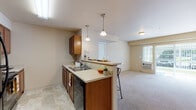 Located at 15913 East 4th Avenue in Spokane Valley, WA, this single-floor unit offers a practical and comfortable layout designed for everyday living. The kitchen is a standout feature, boasting a galley-style design with warm wood cabinetry, light countertops, tile flooring, and black appliances including a range, microwave, and dishwasher. A peninsula with pendant lighting and bar stool seating opens directly to the living area, creating a connected, social atmosphere. The living room is generously sized with soft carpeting, vertical blinds over a sliding door, and a wall-mounted thermostat, contributing to a cozy and functional environment. Natural light filters in through multiple windows fitted with blinds throughout the unit. The bedrooms are carpeted and well-proportioned, each featuring windows that bring in outdoor light and views of the surrounding neighborhood. The bathroom includes wood vanity cabinetry, light countertops, tile flooring, and a shower with a curtain, accompanied by open shelving for storage. A dedicated laundry room with side-by-side washer and dryer hookups and built-in shelving adds significant convenience. The overall aesthetic is neutral and clean, with warm beige tones, consistent finishes, and a well-organized floor plan throughout.