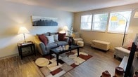 This single-floor space presents a welcoming living area designed with comfort and functionality in mind. The room features warm hardwood-style flooring that extends throughout, providing a cohesive and clean foundation. Neutral-toned walls in a soft beige create a calm, versatile backdrop that complements a variety of decor styles.

A standout feature of the living area is the natural light streaming in through a sizeable window, which offers a pleasant view of outdoor greenery, enhancing the overall brightness and openness of the room. The textured ceiling adds subtle architectural character to the space.

The living area is well-proportioned, offering enough room to accommodate a sofa, accent chair, storage ottoman, and a central coffee table without feeling crowded. A bold geometric area rug in earth tones anchors the seating arrangement, adding visual interest and warmth. A floor lamp and decorative elements, including a skyline wall canvas and floral accents, contribute to the inviting ambiance. The overall atmosphere is relaxed and livable, with practical touches that make the space feel both stylish and functional.