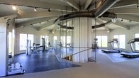 This single-floor fitness facility is designed with both functionality and spaciousness in mind. The soaring vaulted ceiling with exposed white-painted wood beams creates an open, airy atmosphere, while large industrial-style HVAC ducts run overhead, lending the space an bold, utilitarian aesthetic. Track lighting is mounted along the ceiling lines, ensuring even illumination throughout.

At the center of the space stands a striking cylindrical rope climb structure, surrounded by thick climbing ropes that extend from floor to ceiling — a standout functional training feature that serves as the focal point of the room. The flooring transitions between rubberized athletic matting and carpet, delineating different training zones across the generous floor plan.

The perimeter is lined with an extensive selection of strength training machines and benches, providing options for a full-body workout. Cardio equipment, including ellipticals and other machines, is positioned near the windows. Multiple large windows allow abundant natural light to fill the space, offering views of the surrounding outdoor landscape and creating an energizing environment for exercise.

Overall, this fitness space balances professional-grade equipment with an expansive, light-filled design that is both practical and motivating.