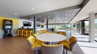 This single-floor amenity space is designed as a dynamic social hub, combining entertainment, dining, and relaxation within one open, interconnected layout. A striking sculptural ceiling panel with a repeating organic pattern serves as a defining architectural feature, adding texture and visual interest throughout the main gathering area. The flooring is a warm-toned hardwood that runs consistently across the space, unifying the various zones.

The central area features round and long rectangular tables paired with bold yellow upholstered chairs, providing flexible seating for casual dining or group gatherings. A bar-style counter with yellow metal stools lines one wall, equipped with mounted flat-screen televisions, wood-faced cabinetry, dark countertops, and beverage appliances. 

A dedicated games zone includes a pool table, shuffleboard table, electronic dartboard, and foosball table, all bathed in natural light from expansive floor-to-ceiling glass doors that open to an outdoor terrace. A separate lounge nook offers a comfortable armchair, patterned area rug, and a large wall-mounted television for quiet viewing. The abundant glazing throughout provides panoramic outdoor views and connects the interior seamlessly to the exterior terrace.
